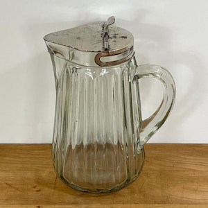 May include: A clear glass pitcher with vertical ribbed detailing and a metal lid. The pitcher has a handle and a spout. The lid is attached with a hinge. The pitcher is sitting on a wooden surface.