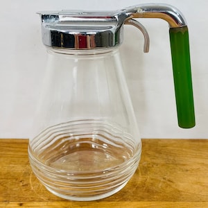 May include: A clear glass syrup pitcher with a chrome lid and handle. The handle is green and the pitcher has a ribbed design around the base. The pitcher is sitting on a wooden surface.