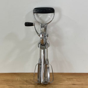 May include: Vintage hand-crank egg beater with a black handle and top grip. The beater is made of shiny, polished metal and features two beaters. The design is a classic kitchen tool.