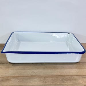 May include: A rectangular white enamel baking dish with a vibrant blue trim. The dish is empty and sits on a wooden surface. The baking dish is a classic kitchenware item.