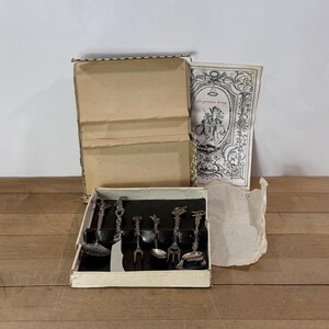 May include: A set of six silver-toned metal utensils with ornate handles arranged in a rectangular cardboard box. The set includes a small ladle, a knife, a fork, and three spoons. A paper insert with black and white illustrations is included.