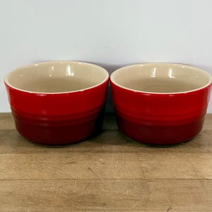 May include: Two red ceramic bowls with a cream-colored interior. The bowls are round with a slightly flared rim and sit on a wooden surface. The bowls are likely for serving or baking.