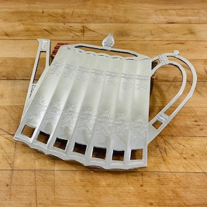 May include: A silver-colored teapot-shaped trivet with a decorative floral pattern. The trivet has a handle and a spout, and is designed to protect surfaces from hot teapots. The trivet measures approximately 10 inches long.