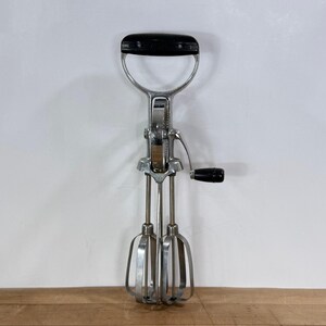 May include: A vintage hand-crank egg beater with a black handle and a chrome-plated finish. The beater features a curved handle, a gear mechanism, and two wire beaters. This kitchen tool is designed for mixing ingredients.