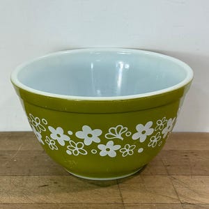 May include: A round mixing bowl with a glossy olive green exterior and a smooth white interior. The outer surface features a repeating white floral pattern consisting of five-petaled flowers, small circles, and stylized leaf shapes encircling the bowl.