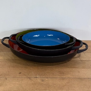 May include: A set of three nested round pans with black exterior surfaces and two side handles each. The largest pan has a red interior, the middle pan has a green interior, and the smallest top pan has a vibrant blue interior surface.