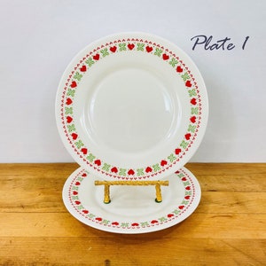May include: Two white ceramic plates with a red heart and green leaf border design. The plates are displayed on a wooden surface. The text "Plate 1" is visible in the upper right corner.