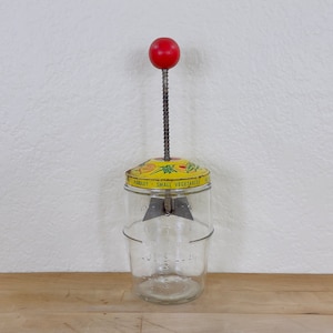 May include: Vintage glass vegetable chopper with a yellow lid featuring the words "PARSLEY SMALL VEGETABLES." It has a red wooden knob and a metal spring. The clear glass jar has measurement markings.