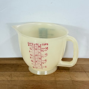 May include: A vintage off-white plastic measuring cup with a handle and a spout. Red markings indicate cup and ounce measurements. The cup is designed for kitchen use and sits on a wooden surface.