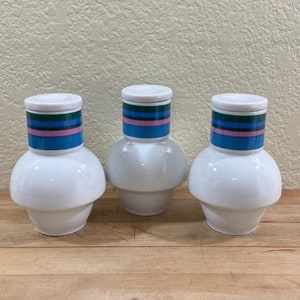 May include: Three white ceramic vases with a bulbous base and a cylindrical neck. Each vase has a white lid and a band of blue, pink, and green stripes around the neck. The vases are arranged on a wooden surface.