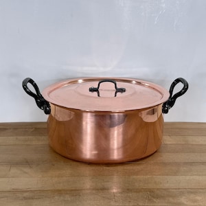 May include: A round copper pot with a matching lid, featuring two black curved side handles and a small black handle on the lid. The pot has a polished metallic finish and a wide, cylindrical body, showcasing a classic kitchen cookware design.