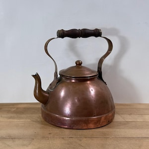 May include: A vintage copper kettle with a rounded body, spout, and lid. The kettle features a curved handle and a wooden grip. The copper has a warm, reddish-brown hue, and the wooden handle is a dark brown. The kettle is placed on a wooden surface.