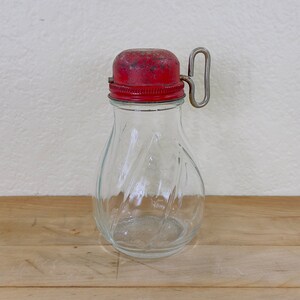May include: A clear glass jar with a swirling ribbed texture and a wide base. The jar features a red metal screw-on lid with a weathered finish and a vertical metal loop handle attached to the side of the cap for easy gripping and pouring.