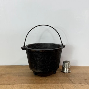 May include: A small, black cast iron cauldron with a rounded body, three short legs, and a thin, arched metal handle. The cauldron is positioned next to a single silver-colored metal thimble, providing a clear sense of the small scale.