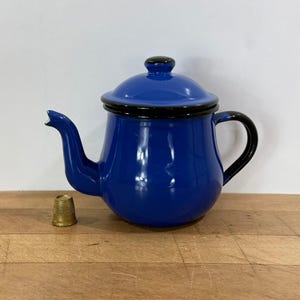 May include: A glossy cobalt blue enamel teapot with a matching lid and a black handle, spout, and rim. A small, textured brass thimble is positioned next to the teapot to provide a sense of scale for the object's overall size.