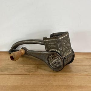 May include: An antique metal food grater with a wooden handle. The grater is a dark gray color and has the words "PATENTS PENDING" and "FOOD GRATER" embossed on the side. The grater is sitting on a wooden surface.