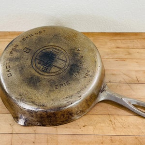 May include: An antique Griswold cast iron skillet, size 8, with a handle. The skillet's underside displays the brand's logo and text, including "ERIE PA U.S.A." and "CAST IRON SKILLET". The pan has a well-worn, aged appearance.
