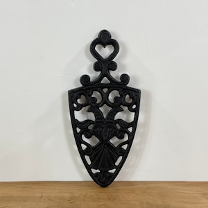 May include: Black cast iron trivet with an ornate, openwork design. The trivet features a heart-shaped top and a shield-like body with floral and scroll details. It is designed to protect surfaces from hot cookware.