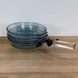 May include: A stack of three blue-tinted glass baking dishes with a black handle and a clear glass handle. The dishes are round and have a slightly flared rim. The handle is attached to the side of the bottom dish.