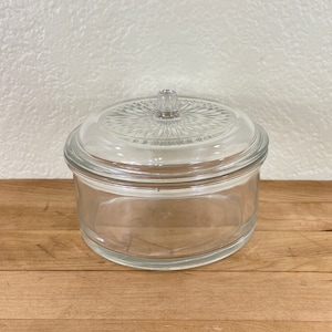 May include: A clear glass round container with a matching lid. The lid has a decorative pattern and a small knob. The container sits on a wooden surface.