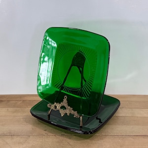 May include: Two square, emerald green glass plates with a decorative design in the center. The plates are displayed on a gold-colored metal stand. The plates are sitting on a wooden surface.