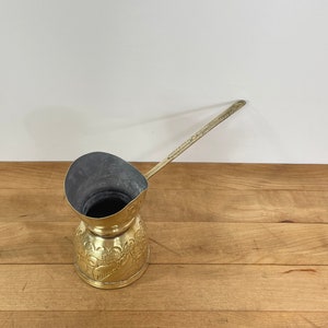 May include: A vintage brass coffee pot with a long handle and a decorative floral pattern. The pot has a wide mouth and a dark interior. It sits on a wooden surface against a white background.