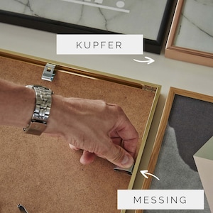 Op de afbeelding: Een close-up van iemand die een metalen ophangclip voor foto's aan een gouden fotolijst bevestigt. De lijst is gelabeld "MESSING" en staat naast een zwarte fotolijst met het label "KUPFER".