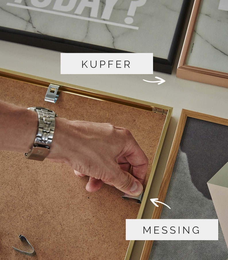 Op de afbeelding: Een close-up van iemand die een metalen ophangclip voor foto's aan een gouden fotolijst bevestigt. De lijst is gelabeld "MESSING" en staat naast een zwarte fotolijst met het label "KUPFER".