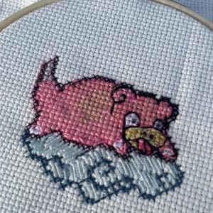 May include: Cross-stitch embroidery of a pink Pokémon character, Snorlax, lying on its back with its tongue sticking out. The character is stitched on white Aida cloth and is surrounded by a blue and white background.