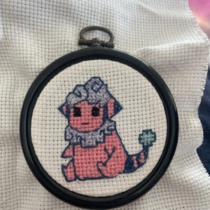 May include: A pink and purple cross-stitch embroidery of a cartoon animal with a blue flower on its tail, framed in a black circular hoop.