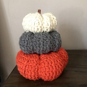 Puede incluir: Una pila de tres calabazas de ganchillo en colores otoñales. La calabaza inferior es naranja, la del medio es gris y la superior es color crema. Un pequeño palito de canela sirve como tallo.