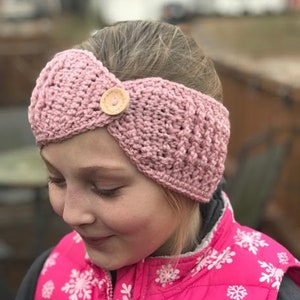 May include: A pink crocheted headband with a wooden button. The headband is worn by a person with a pink vest with a snowflake pattern.