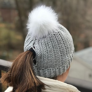 Può includere: Berretto di lana grigio con un pon pon bianco e un foro sul retro per una coda di cavallo.