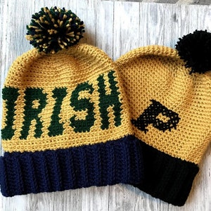 Op de afbeelding: Twee gebreide mutsen met pompoenen. Eén muts is geel met groene letters die "Irish" zeggen en een donkerblauwe rand. De andere muts is geel met een zwart ontwerp en een zwarte rand.