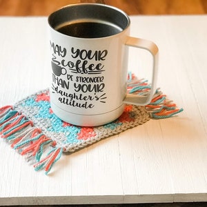 May include: White coffee mug with the text "May your coffee be stronger than your daughter's attitude" on a crocheted coaster with pink and blue fringe.
