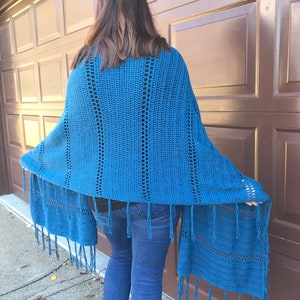 Puede incluir: Un chal de ganchillo azul con flecos. El chal está drapeado sobre los hombros de una persona y los flecos cuelgan.