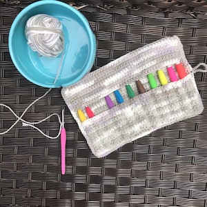 May include: A crocheted gray and white pouch with a drawstring closure. The pouch is filled with colorful crochet hooks. The pouch is sitting on a brown wicker surface.