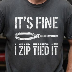 May include: A dark gray t-shirt with the text "IT'S FINE I ZIP TIED IT" in white. The shirt features a graphic of a zip tie. The person in the image is in a garage setting.