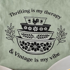 Puede incluir: Camiseta verde salvia con el texto "Thrifting is my therapy & Vintage is my vibe" y un gráfico de cuencos vintage apilados. Los cuencos son negros y blancos con diseños florales y de gallos. La imagen incluye cuencos vintage.