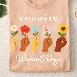 Puede incluir: Camiseta color melocotón con el texto "Happy International Women's Day". La camiseta presenta ilustraciones de manos sosteniendo varias flores: una margarita, un hibisco, una flor azul, un lirio y un girasol. También se ven una taza de café y un pin.