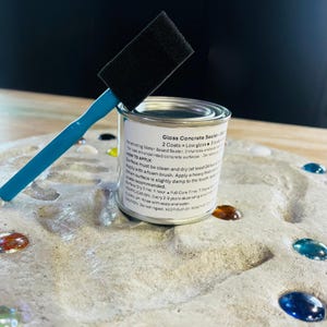 May include: A small metal can of "Gloss Concrete Sealer" with a black foam brush and a blue handle. The can has a white label with black text. The brush is resting on top of the can. The can is on a gray, textured surface with colorful glass beads.