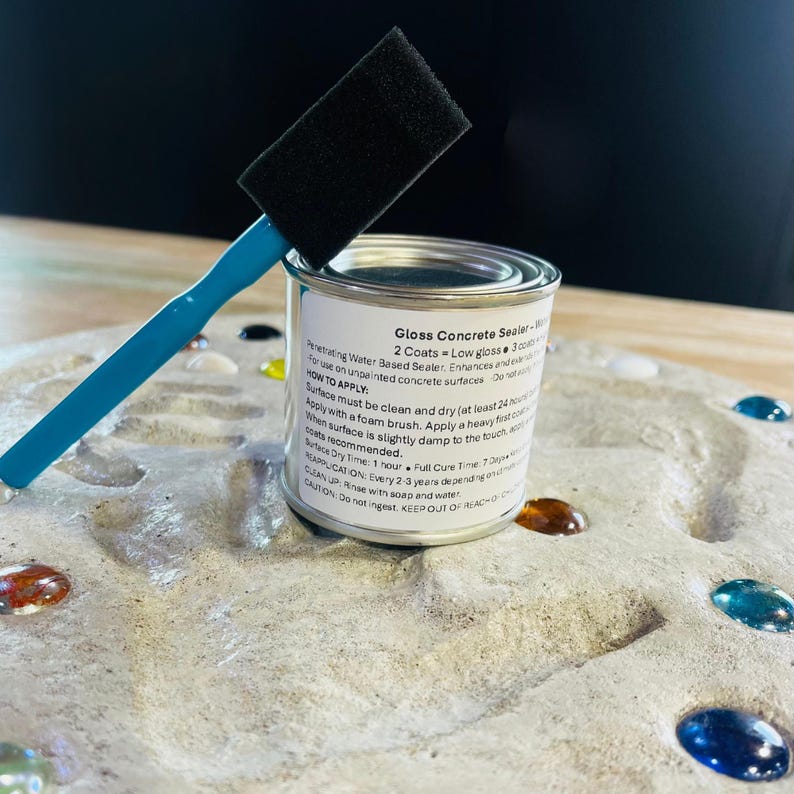May include: A small metal can of "Gloss Concrete Sealer" with a black foam brush and a blue handle. The can has a white label with black text. The brush is resting on top of the can. The can is on a gray, textured surface with colorful glass beads.