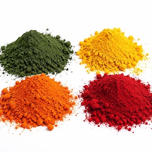 May include: Four distinct mounds of fine, powdery pigment arranged in a square formation. The colors include a deep forest green, a vibrant golden yellow, a bright orange, and a bold, saturated red, each forming a conical shape on a white surface.