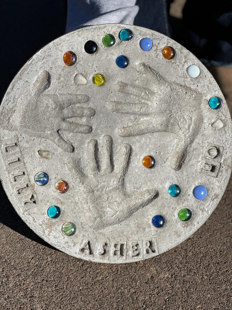 May include: A round, gray concrete stepping stone with handprints and colorful glass gems. The names "LILLY" and "ASHER" are imprinted on the stone. The gems are various colors, including blue, green, orange, and white.