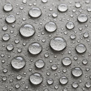 May include: Close-up of clear water droplets of varying sizes on a gray, textured surface. The droplets reflect light, creating a glistening effect. The surface appears to be stone or concrete, with a rough, uneven texture.