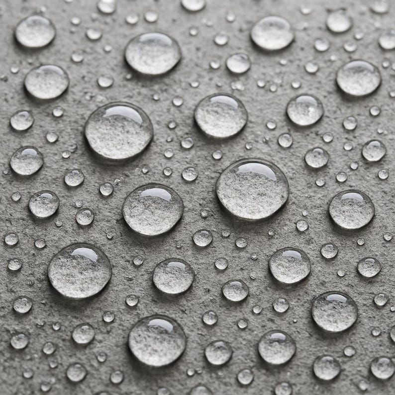 May include: Close-up of clear water droplets of varying sizes on a gray, textured surface. The droplets reflect light, creating a glistening effect. The surface appears to be stone or concrete, with a rough, uneven texture.