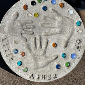 May include: A round, gray concrete stepping stone with handprints and colorful glass gems. The names "Lilly" and "Asher" are inscribed. The stone is decorated with various colored glass marbles.