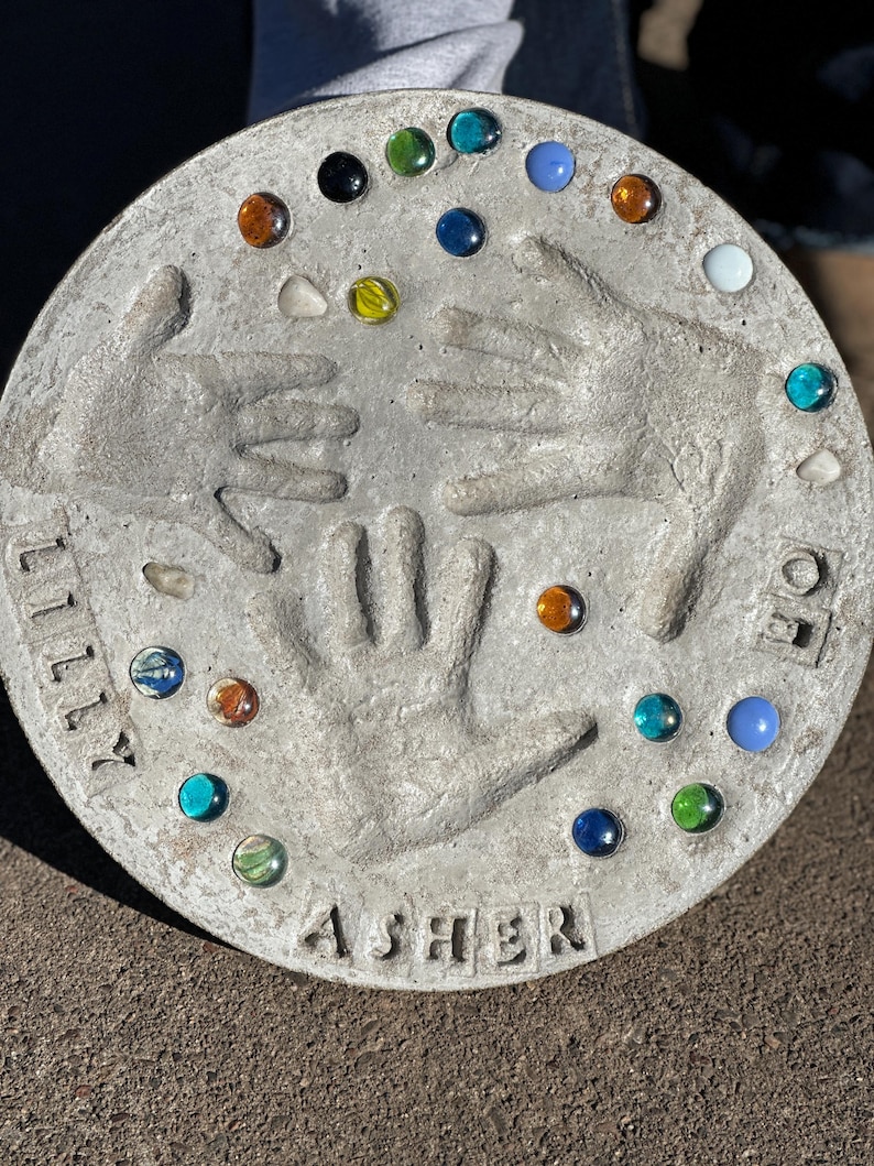May include: A round, gray concrete stepping stone with handprints and colorful glass gems. The names "Lilly" and "Asher" are inscribed. The stone is decorated with various colored glass marbles.