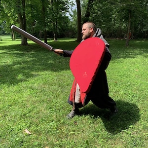 May include: A person in a black hooded cloak and boots practices with a red shield and a gray padded weapon in a grassy area with trees. The shield is oval-shaped.