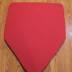 May include: A bright red, pentagon-shaped baseball base. The base is made of a textured material and is set against a wooden floor.